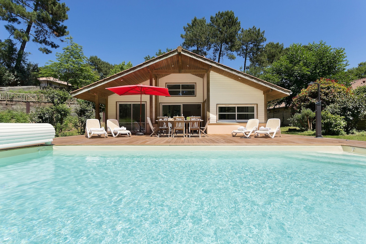 A spacious villa is set against a clear blue sky, showcasing a private pool surrounded by sun loungers. A large covered terrace with a dining table is visible, complemented by a red umbrella. Lush greenery and trees frame the property, enhancing the tranquil atmosphere.