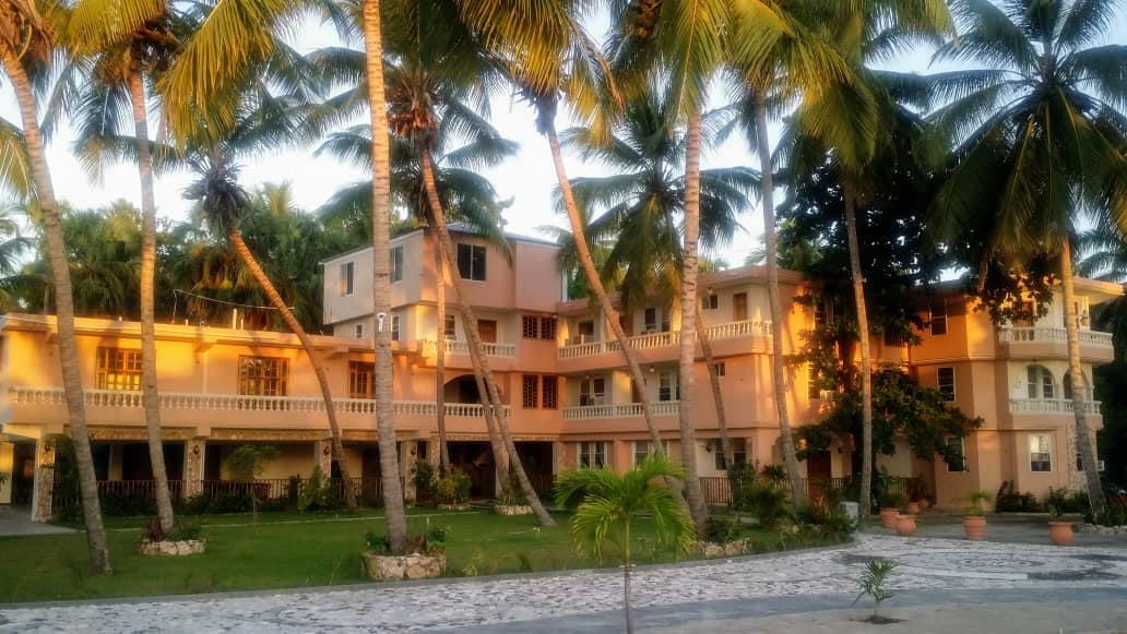 A charming multi-level building is surrounded by tall palm trees and lush greenery, offering a tropical ambiance. The structure features a warm color palette with balconies and terraces, enhancing the outdoor living space. A stone pathway leads through the garden area, connecting to the main entrance.
