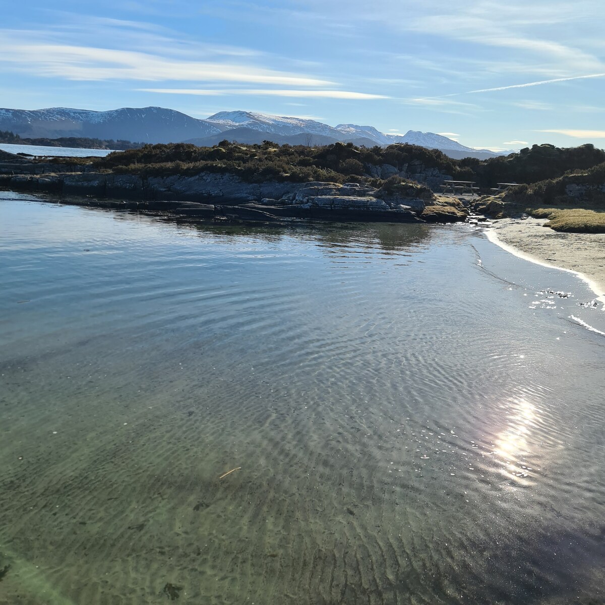 A serene coastal landscape is displayed, featuring gentle waves lapping against a sandy shore. The clear water reflects sunlight, creating shimmering patterns. In the background, a range of mountains is visible, partially covered in snow under a blue sky.