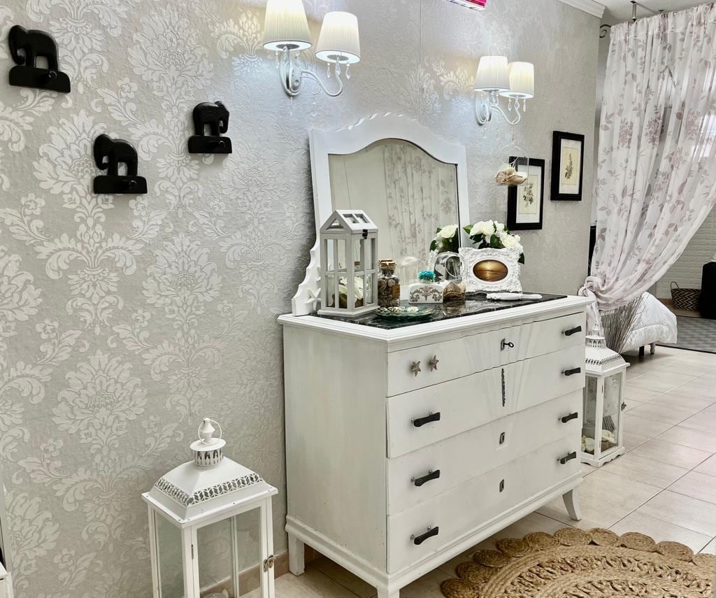 A white dresser stands against a softly patterned wall, complemented by elegant wall sconces providing soft lighting. A mirror reflects decorative items, including a lantern and small plants. Subtle curtains frame a nearby window, enhancing the serene ambiance of the space.