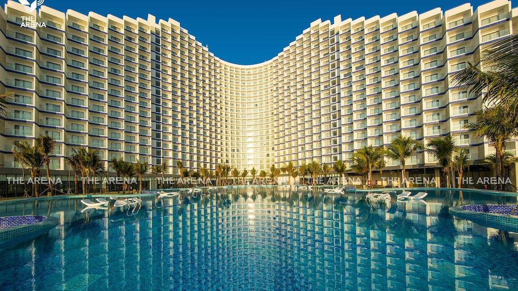A large, modern hotel structure features numerous balconies overlooking a serene swimming pool. Sun loungers and palm trees are positioned around the pool area, reflecting a bright and inviting atmosphere. The building stands tall against a clear blue sky.