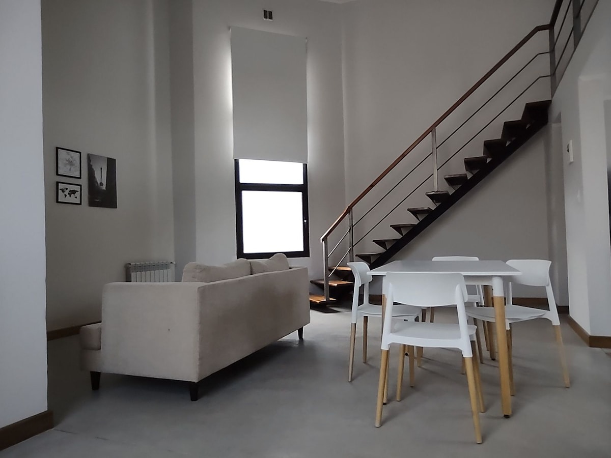 A spacious living area is enhanced by natural light coming through the large window. A beige sofa is positioned adjacent to a white dining table and four chairs. A modern staircase leads to an upper level, with light walls complementing the minimalist design.