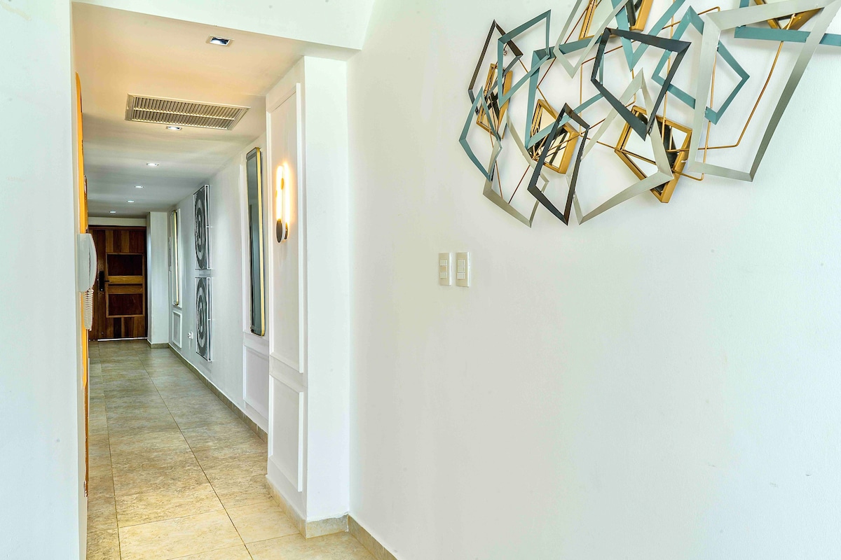 A bright hallway features smooth tile flooring and whitewashed walls. Decorative frames in gold and teal are mounted on the wall, offering a modern artistic touch. Natural light is allowed to flow through windows, contributing to the airy ambiance.