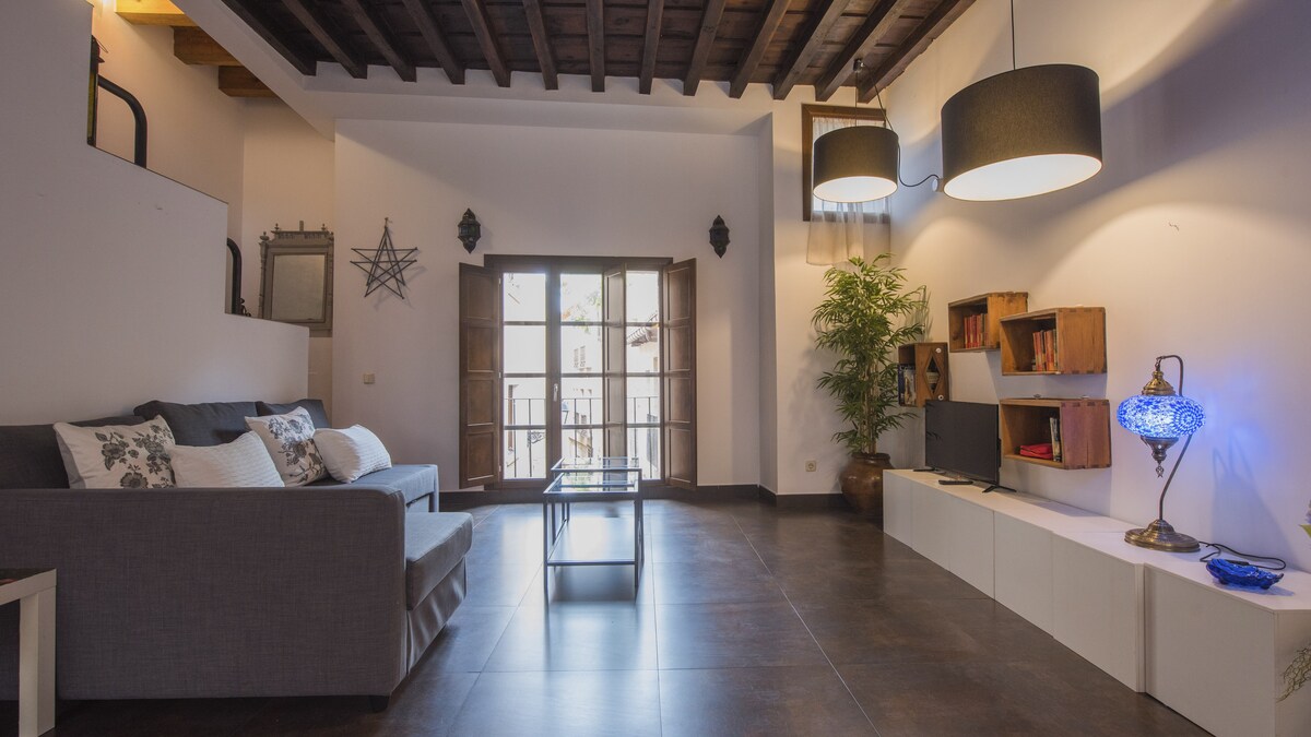 A spacious living area features a comfortable sofa positioned beside large double doors leading to a balcony. Light fixtures hang from a wooden ceiling, illuminating the room. Shelves display decorative items, and a modern television is mounted on the wall above a minimalist console.