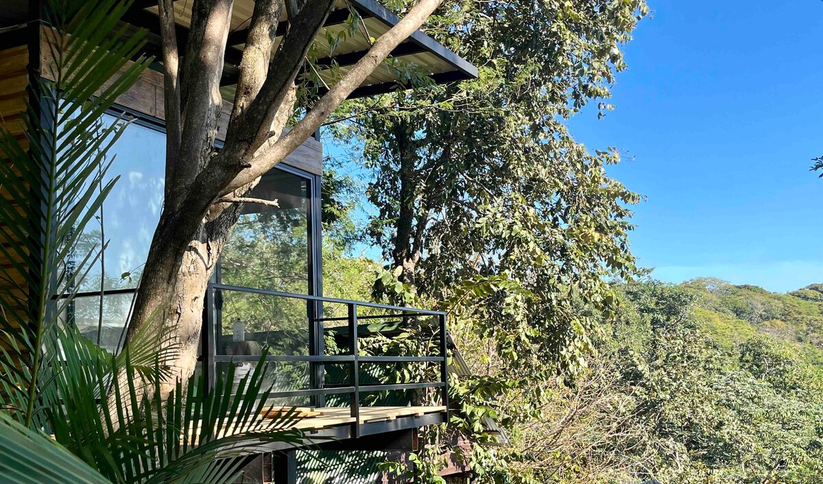 A modern bedroom constructed with glass walls is positioned among trees, providing views of the surrounding nature. A small balcony extends outward, offering an outdoor space to enjoy the scenery. Lush greenery and distant hills contribute to a serene environment.