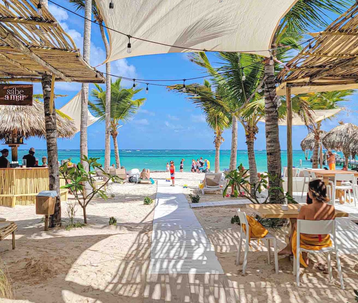 A beach setting features sandy pathways leading towards the water, framed by palm trees and beach cabanas. Tables and chairs are arranged for relaxation. The vibrant ocean horizon is visible, with guests enjoying the sun and beach activities in the background.
