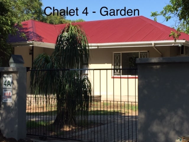 A well-maintained chalet with a red metal roof is framed by lush greenery, featuring a gated entrance. The exterior showcases a light-colored facade and decorative windows, offering an appealing view of the surrounding garden space.