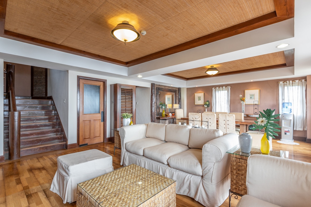 A spacious living area features natural wood finishes and bamboo accents. A comfortable light-colored sofa is positioned near a woven coffee table. Adjacent are elegant armchairs and a dining area. Large windows allow natural light to illuminate the space, enhancing the welcoming ambiance.