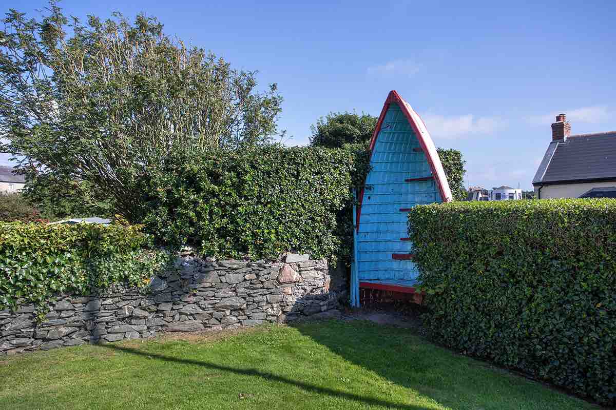 A uniquely designed entrance resembling a boat is framed by lush greenery and a stone wall. The vibrant blue color of the structure contrasts with the surrounding greenery, creating an inviting focal point in the landscaped garden.
