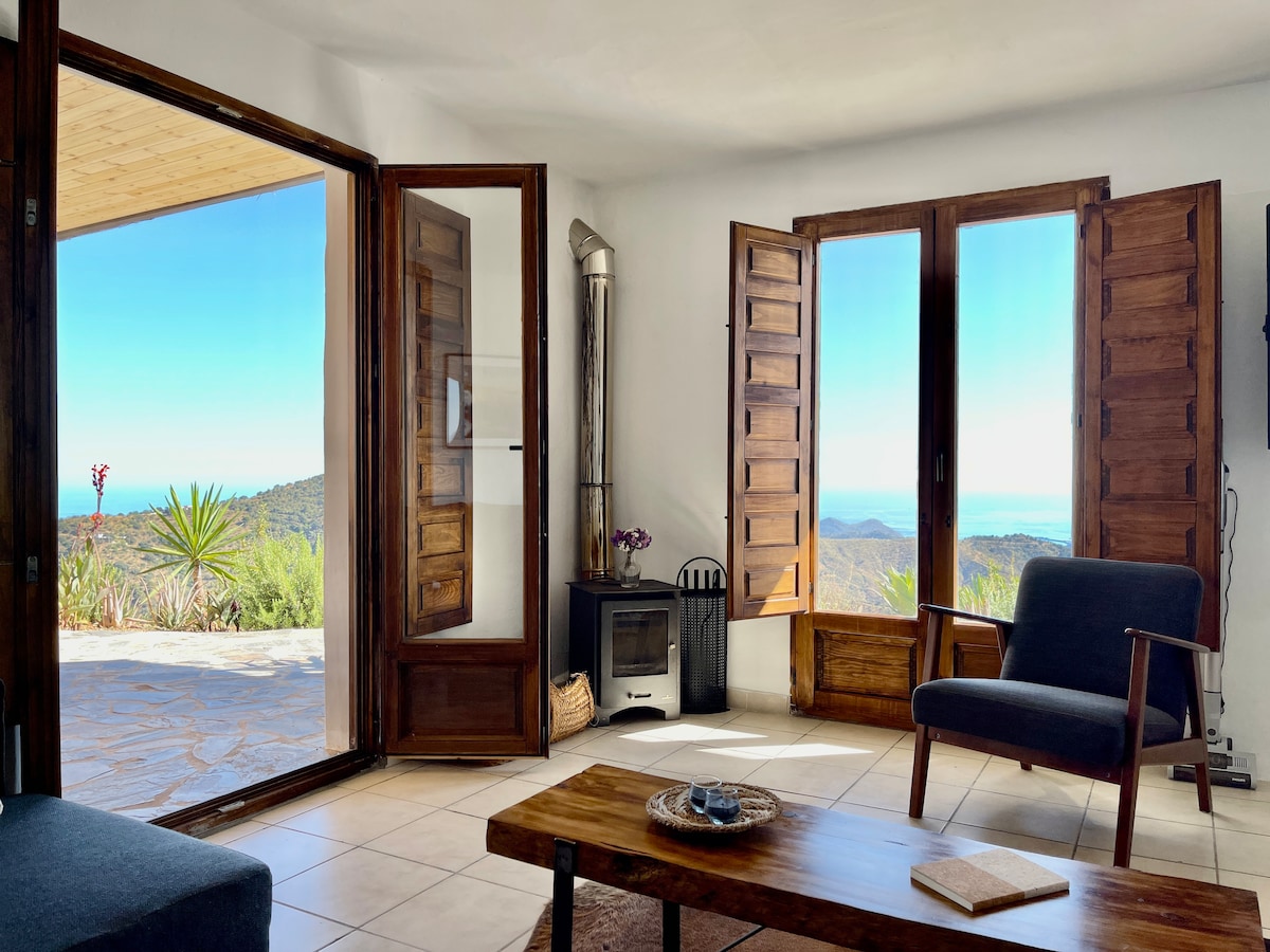 A bright living area features large wooden windows that open to scenic views of the bay. A cozy armchair is positioned near a rustic coffee table, with a wood-burning stove visible in the corner. Natural light floods the space, enhancing its serene ambiance.