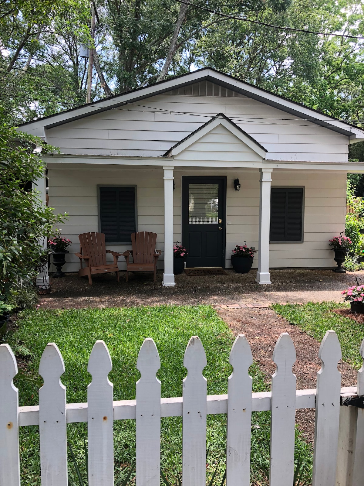 A welcoming front porch features two wooden chairs placed beside a small table, with decorative flower pots enhancing the entrance. The charming exterior is framed by a white picket fence, and lush greenery surrounds the house, creating a serene and inviting environment.