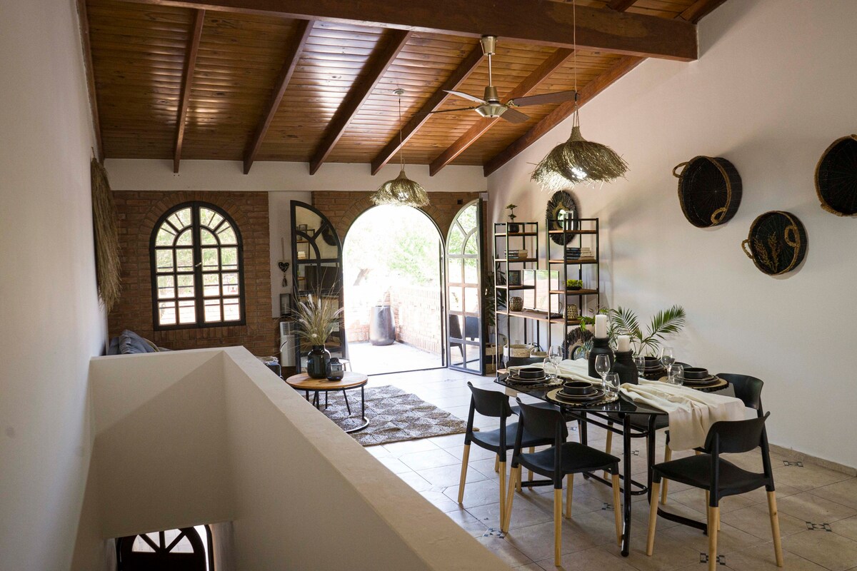A dining area features a spacious table set for six, complemented by black chairs. Large arched windows allow natural light to fill the room, highlighting the wooden ceiling and woven decor. A bookshelf with decorative plants is visible, adding a touch of greenery.
