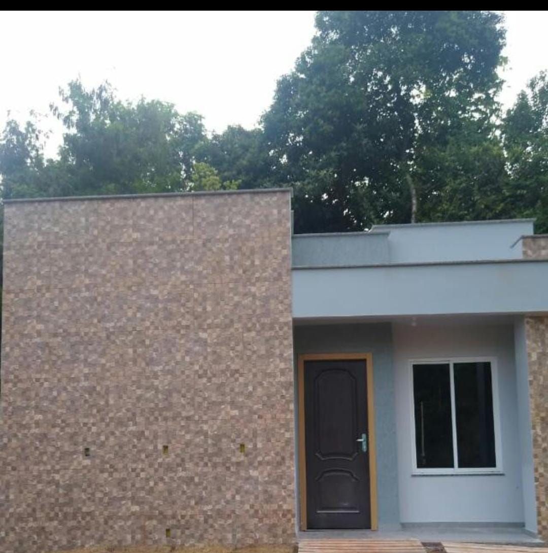The exterior features a modern building facade with a combination of textured stone and smooth grey surfaces. A dark wooden door serves as the main entrance, and a large window allows natural light to fill the interior. Lush greenery surrounds the building.