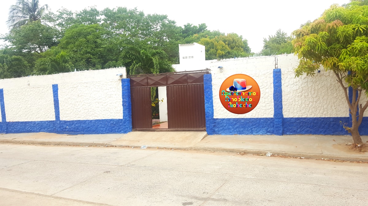 A charming entrance gate is framed by a white and blue wall, surrounded by lush greenery. The gate stands partially open, inviting guests into the property. A colorful sign displaying the property name enhances the welcoming aspect of the entrance.