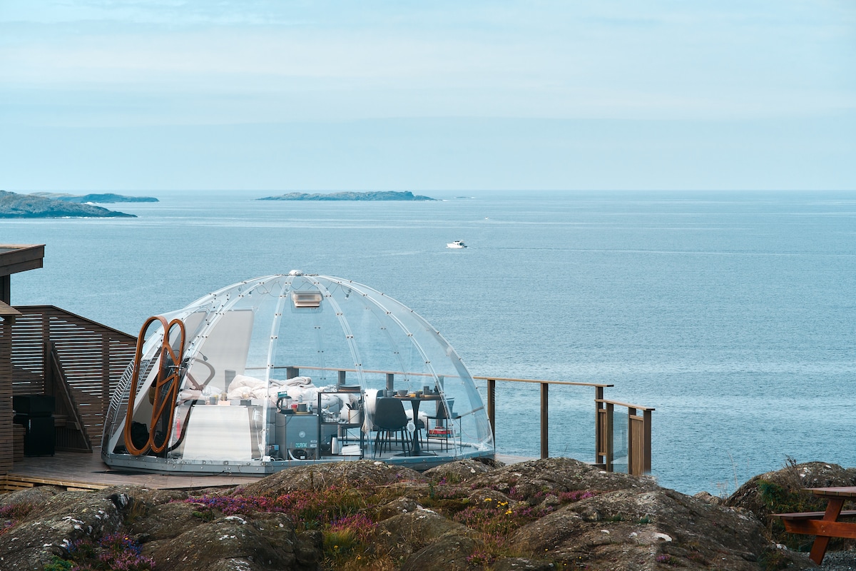 A clear, dome-shaped structure is positioned on rocky terrain, overlooking a vast expanse of the ocean. The interior is visible, featuring a dining area with tables and chairs. The horizon showcases gentle waves and distant islands under a serene sky.