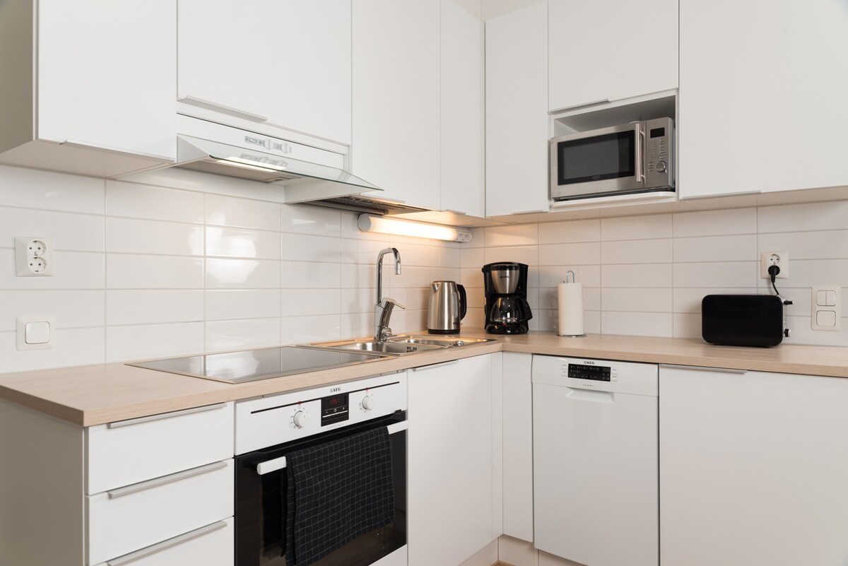 A modern kitchen is presented with sleek white cabinetry and a light countertop. Essential appliances, including a microwave, coffee maker, and dishwasher, are visible. A stainless steel sink is positioned under a wall-mounted light for ample illumination.