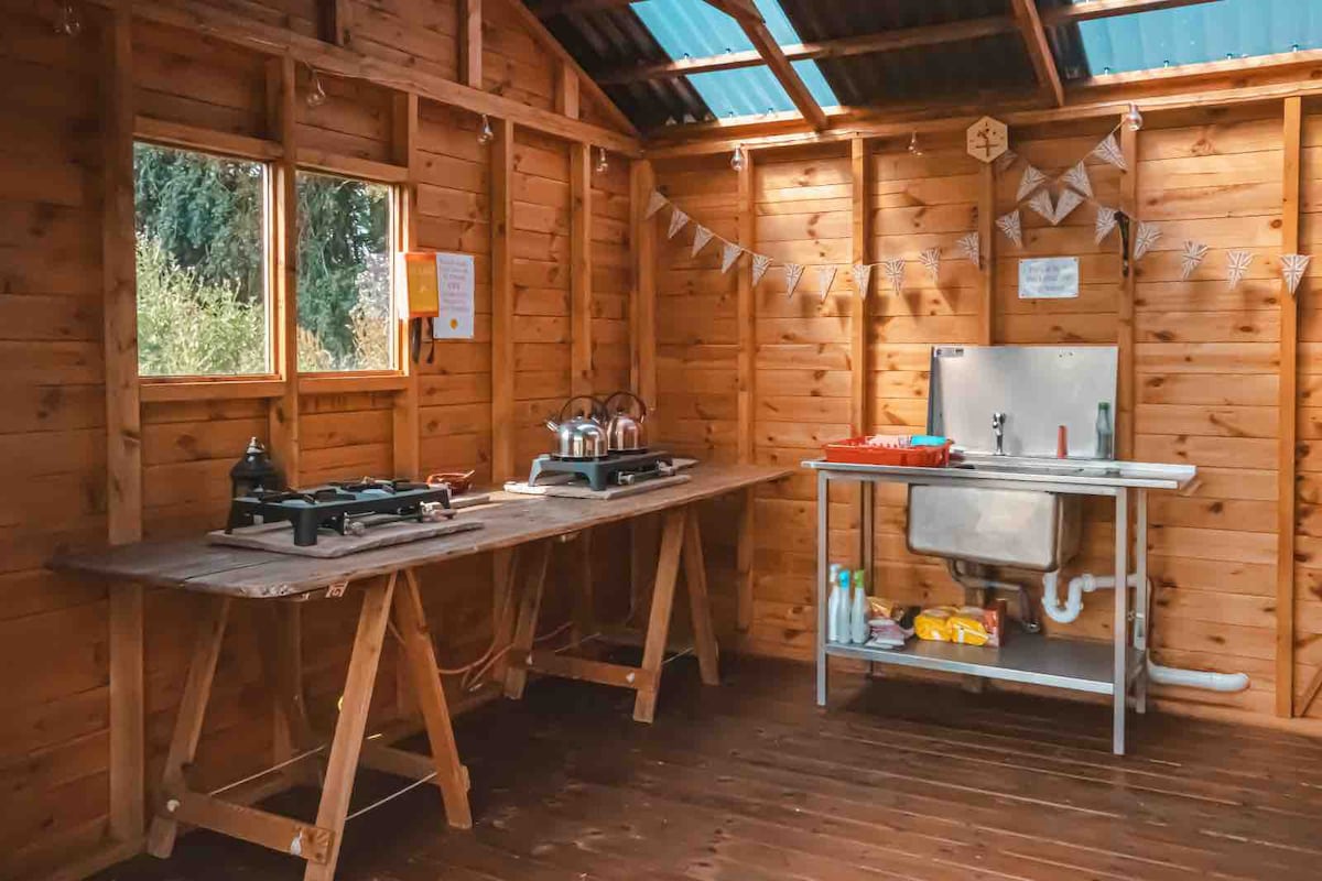 A communal field kitchen features wooden walls and a wooden floor, illuminated by natural light from multiple windows. Cooking stations are equipped with gas burners on sturdy tables, and a stainless steel sink with a countertop is positioned against the wall, complemented by storage items below.