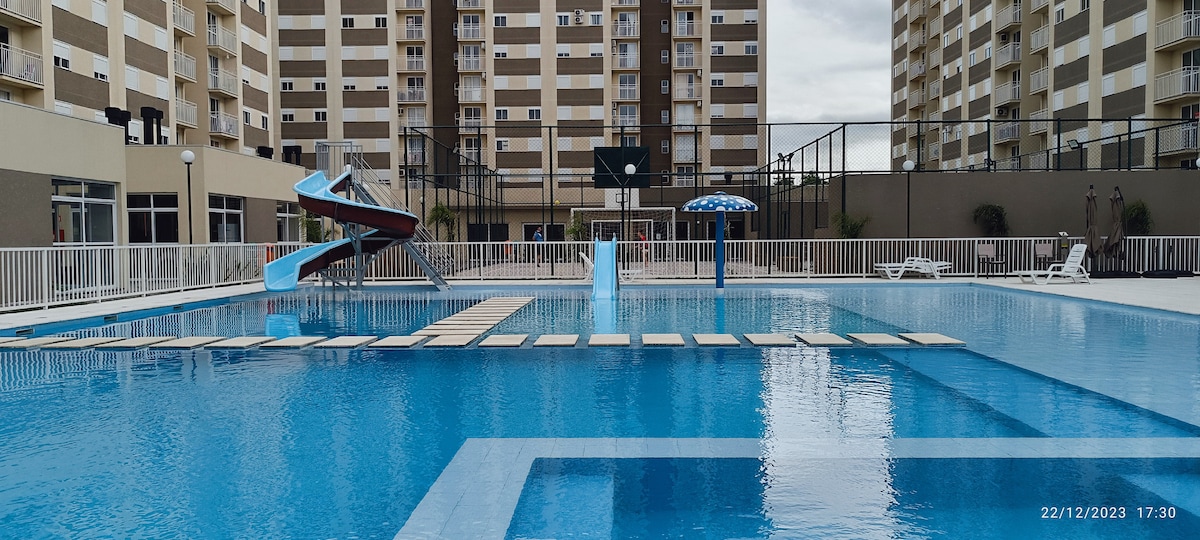 The outdoor pool area features a bright blue pool with a water slide, surrounded by lounge chairs and umbrellas. A basketball court can be seen in the background, along with safety railings along the pool's perimeter.