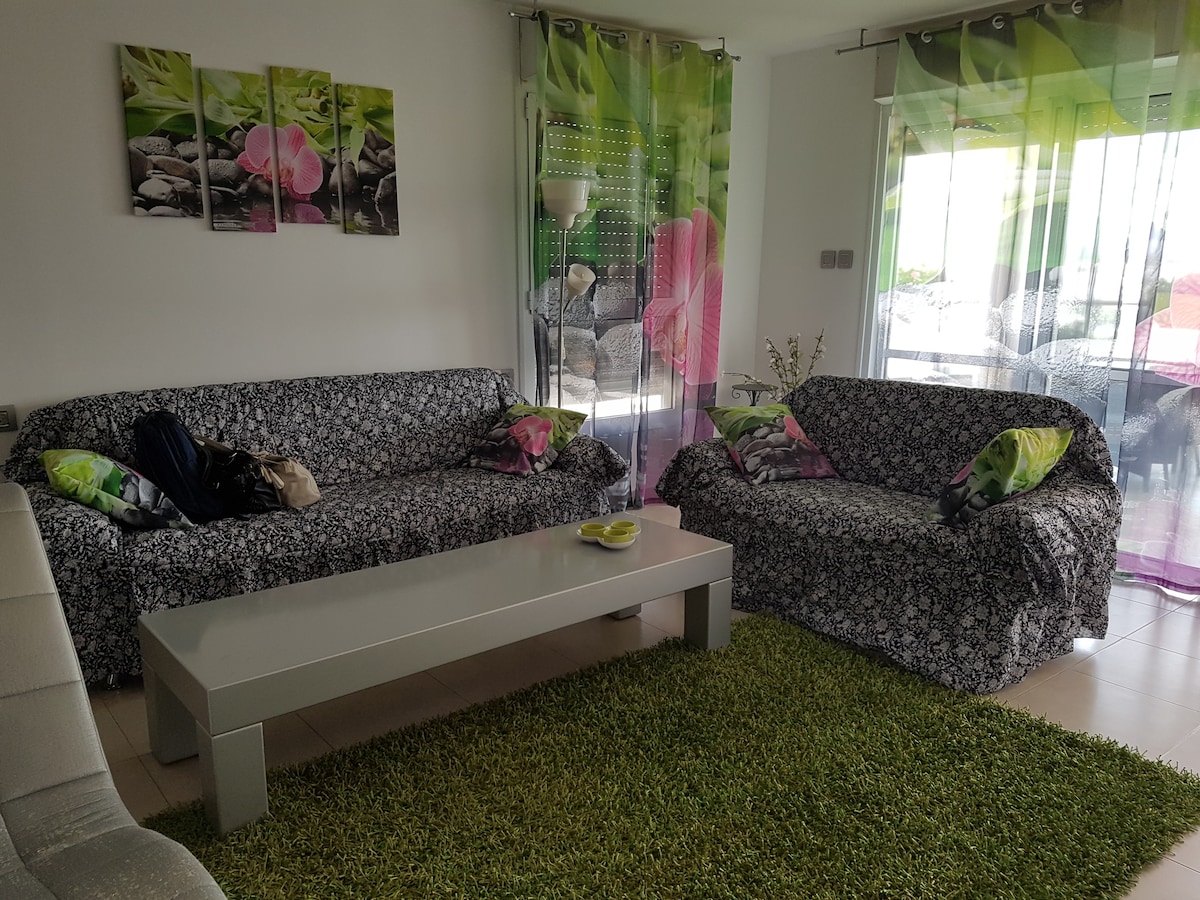 A living area is shown with two floral-patterned sofas and a low coffee table at the center. A vibrant green rug covers the floor, while large glass doors lead to a balcony, allowing natural light to enhance the space. Colorful curtain panels add a decorative touch.
