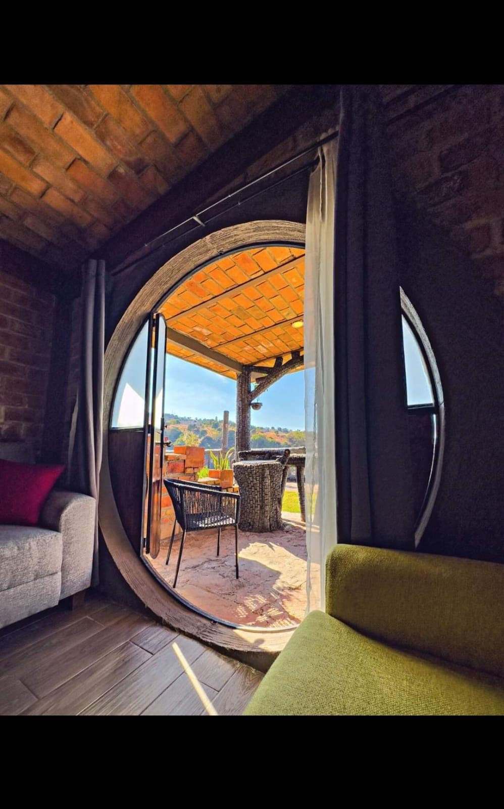 An oval door frames a view of a covered terrace with wicker furniture and a wooden ceiling. Natural light illuminates the interior area featuring a gray couch and a green armchair, while the outdoors offers a glimpse of lush greenery.