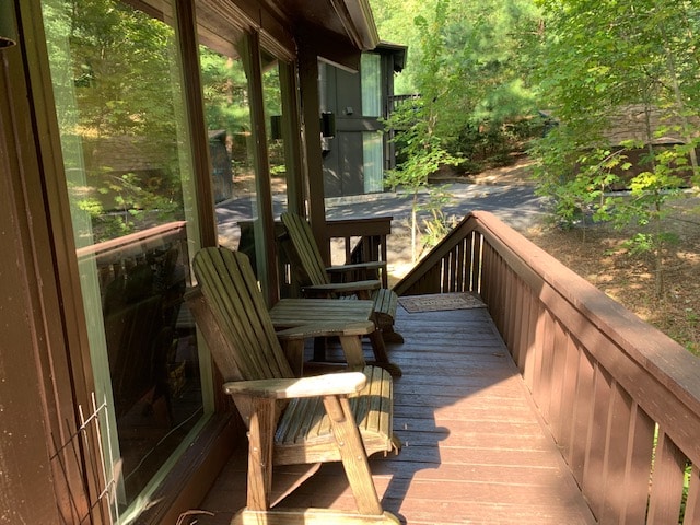 A wooden deck features two Adirondack chairs positioned for enjoying the natural surroundings. Large windows provide a glimpse of the lush greenery outside, while the peaceful path is visible in the background, enhancing the tranquil outdoor space.