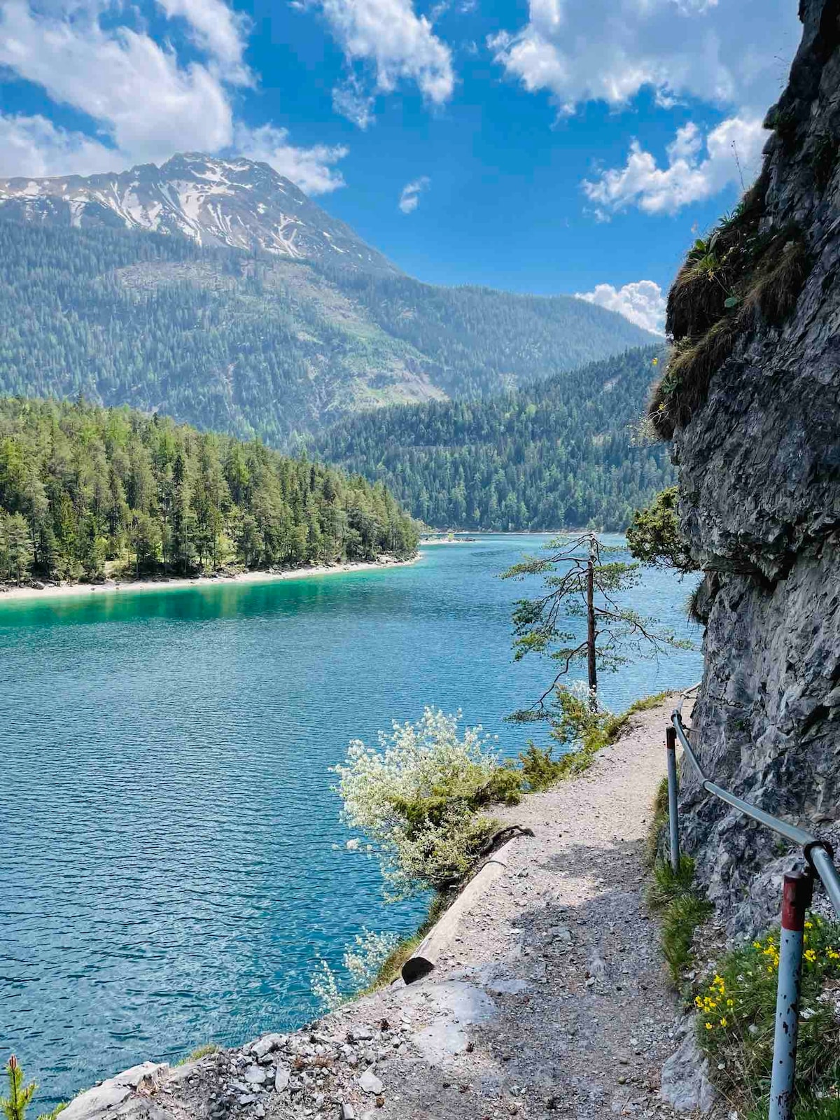 A scenic view of a tranquil lake surrounded by lush green trees and mountains in the background. Sunlight reflects off the water, creating a serene blue hue. A narrow pathway alongside the lake is visible, with a guardrail ensuring safety.