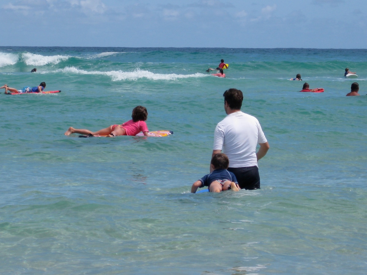 A clear, blue ocean scene features several swimmers enjoying the water. A parent is seen assisting a child on a boogie board, while other beachgoers surf and float in the waves, all under a bright sky dotted with clouds.