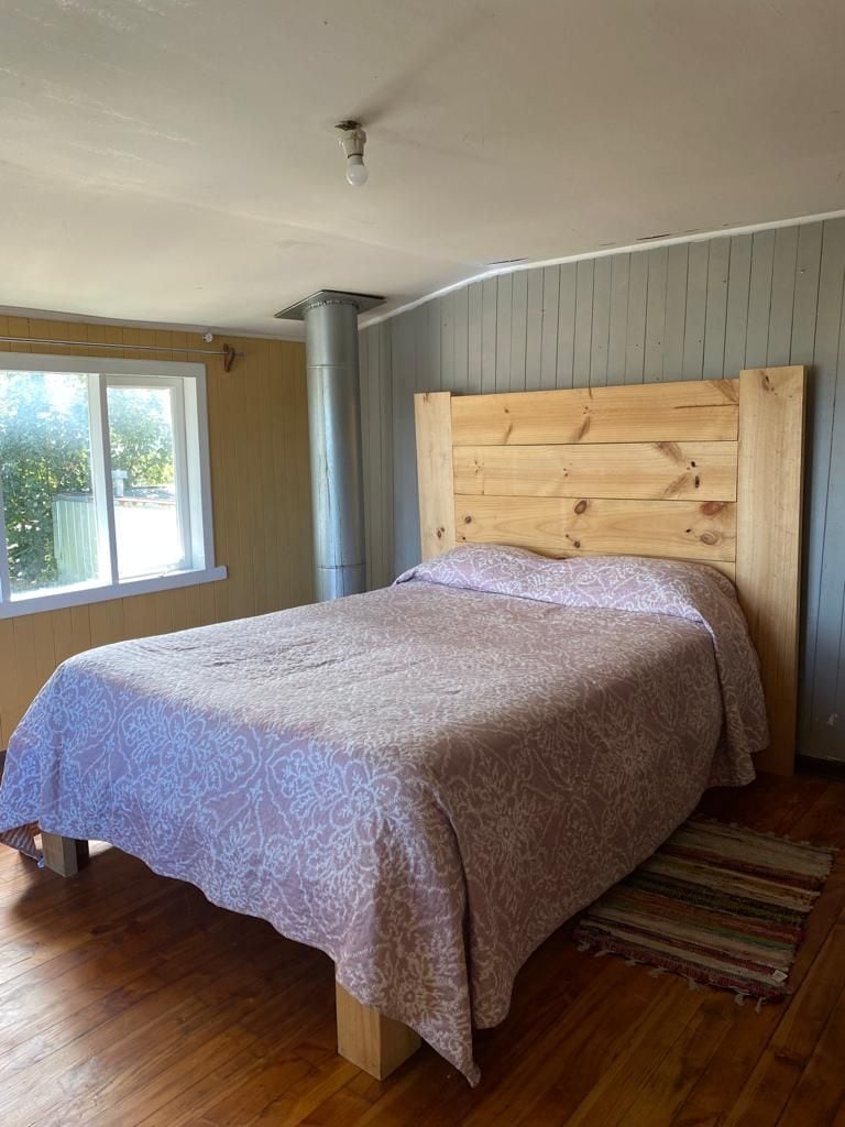 A spacious sleeping area features a wooden bed frame with a patterned blanket. Natural light enters through a nearby window, illuminating the warm wood floors and lightly colored walls. A cozy rug adds texture to the room, enhancing the inviting atmosphere.