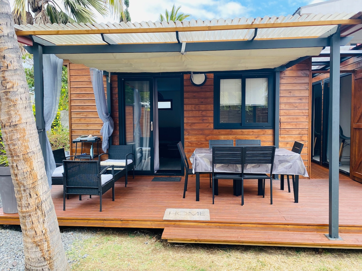 A covered outdoor terrace is seen with a wooden deck and seating areas. Black wicker chairs and a dining table are arranged to one side, while the entrance to the bungalow is visible through sliding glass doors. Lush greenery surrounds the space.
