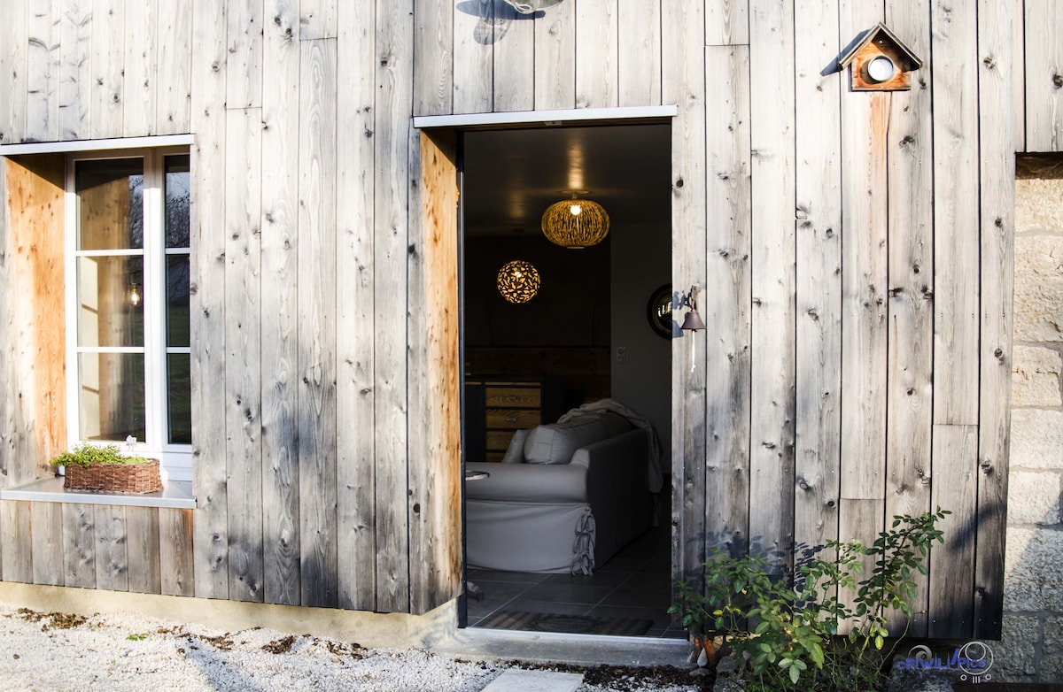 The entrance to the studio is framed by rustic wooden panels, with a door opened to reveal a glimpse of the cozy interior. A decorative light fixture hangs above, and a nearby window showcases natural light illuminating the space.