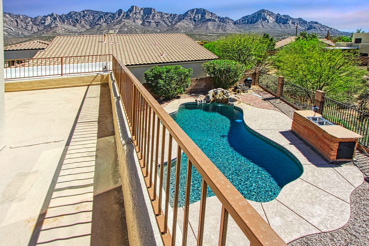 An aerial view showcases a serene backyard with a sparkling pool shaped in a gentle curve, surrounded by paved patio and lush greenery. The striking mountain range is visible in the background, creating a scenic backdrop against a clear sky.