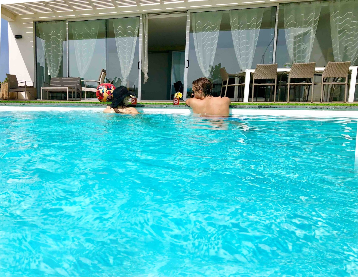 A heated pool is visible in the foreground, with two individuals enjoying the water. Lounge chairs are arranged nearby, and expansive glass doors can be seen in the background, providing access to a shaded outdoor area.