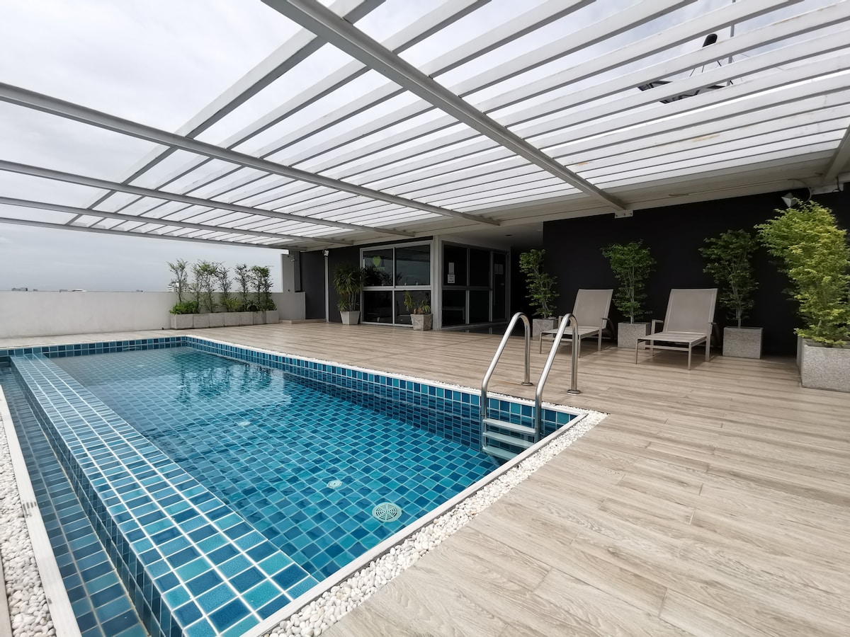 A rooftop area is shown featuring a small swimming pool, surrounded by a sun deck with two lounge chairs. Potted plants are placed along the perimeter, and large windows provide a view of the interior space beyond.