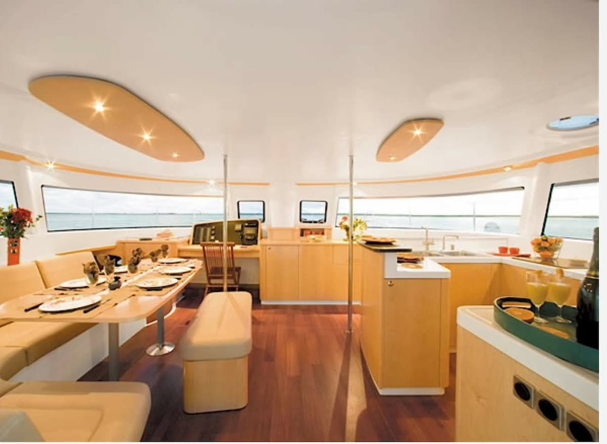 A spacious interior of the catamaran features warm wooden accents and modern amenities. The dining area is equipped with a long table surrounded by comfortable seating, while large windows allow natural light to illuminate the space. A well-equipped kitchen area is visible, contributing to the functional layout.