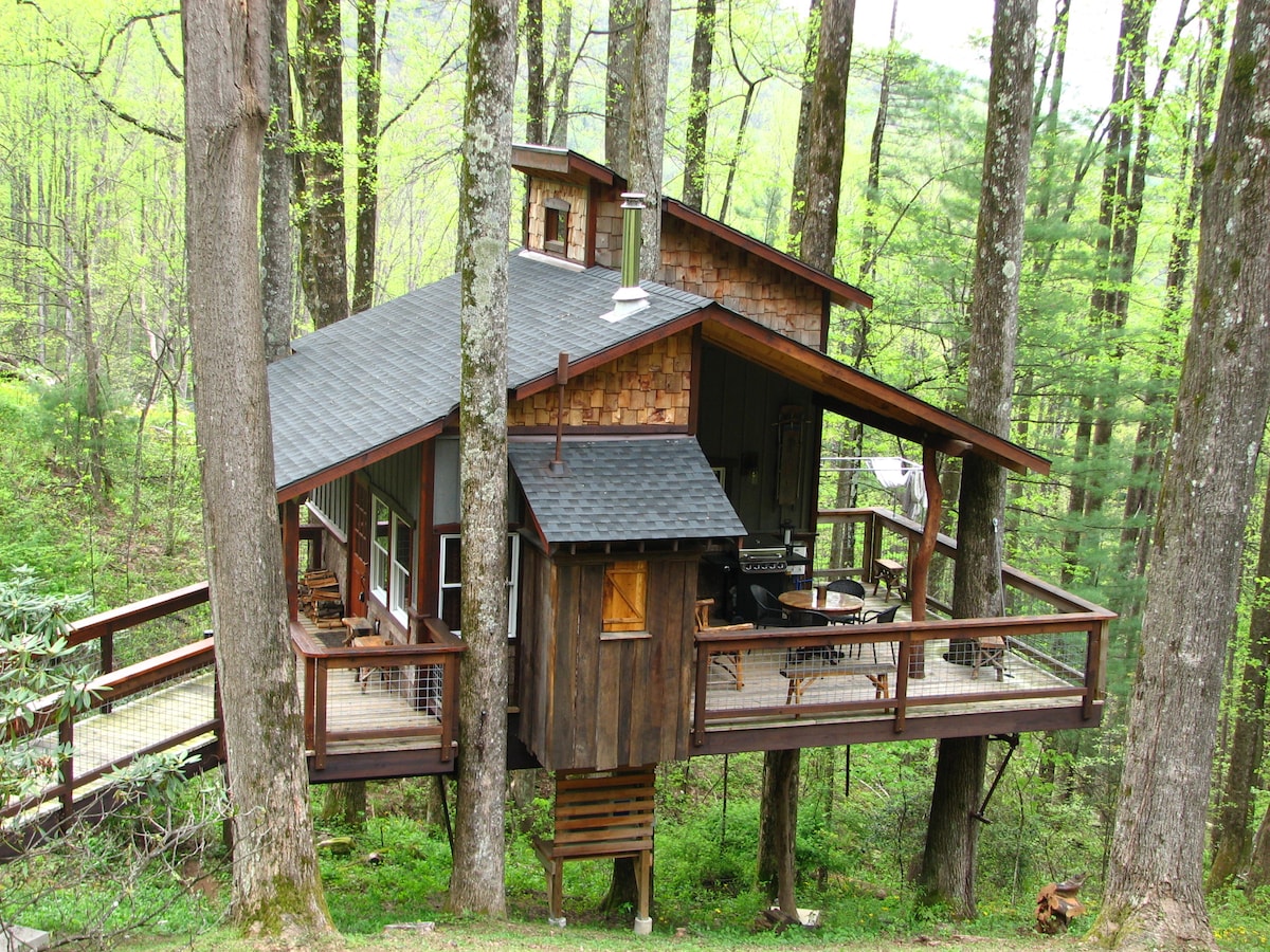 A treehouse cabin is nestled among tall trees, featuring a spacious deck that extends outward. The structure is built with natural materials, and a grill is visible on the deck. Lush green foliage surrounds the treehouse, enhancing the natural ambiance.