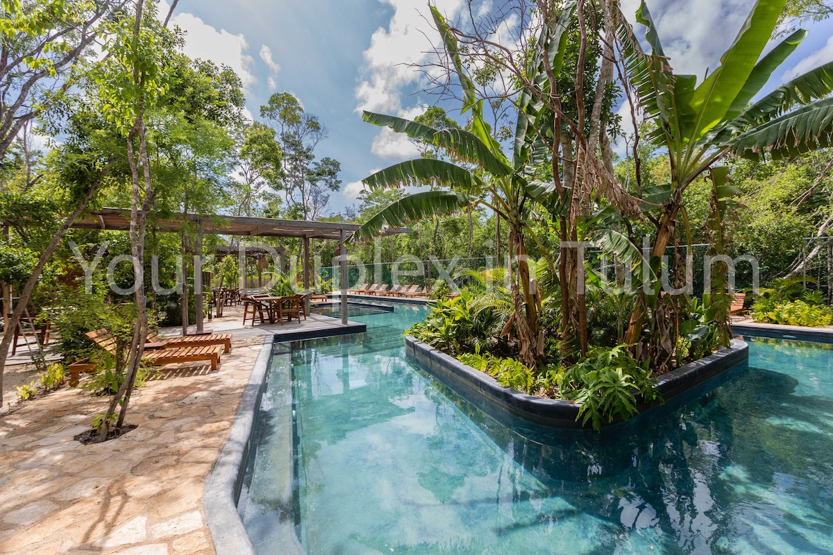 A serene pool area features clear turquoise waters surrounded by lush greenery and tropical plants. Wooden seating areas are visible nearby, offering shaded spots for relaxation. The design integrates nature with the water, creating a tranquil environment for leisure.
