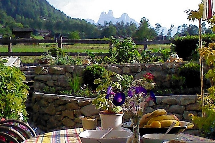 A spacious terrace is visible, adorned with vibrant flowers in pots and a table set with bowls of fruit. The backdrop features expansive Alpine meadows and majestic peaks of the Dolomite range, providing a serene and picturesque setting.