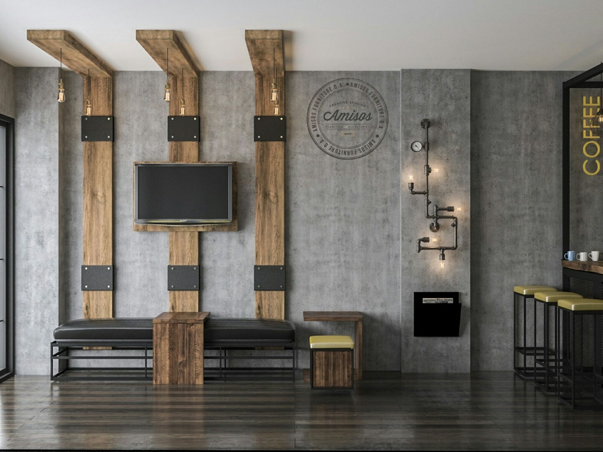 A modern seating area features wooden accents and industrial-style lighting. A wall-mounted television is placed above a sleek bench. A small table complements the seating, while a minimalist coffee bar with tall stools is set against the gray wall.