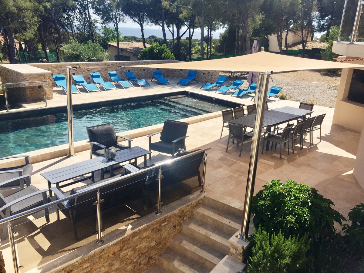 An outdoor area featuring a heated swimming pool surrounded by lounge chairs. A spacious dining table with multiple chairs is situated beside the pool, offering a relaxed space for meals. Lush greenery and shaded spots enhance the tranquil setting, with views of the nearby sea.