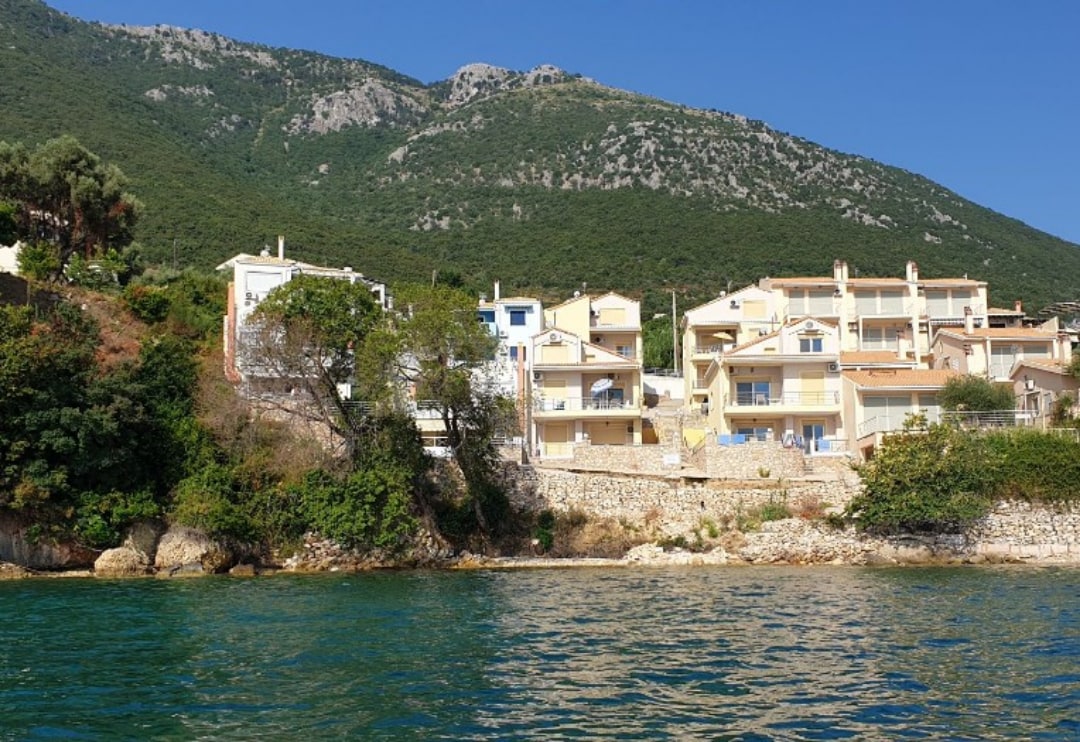 A waterfront view features a cluster of residential buildings set against a backdrop of rolling green hills. The clear blue water reflects a sunny sky, highlighting the natural surroundings and peaceful ambiance of the area.