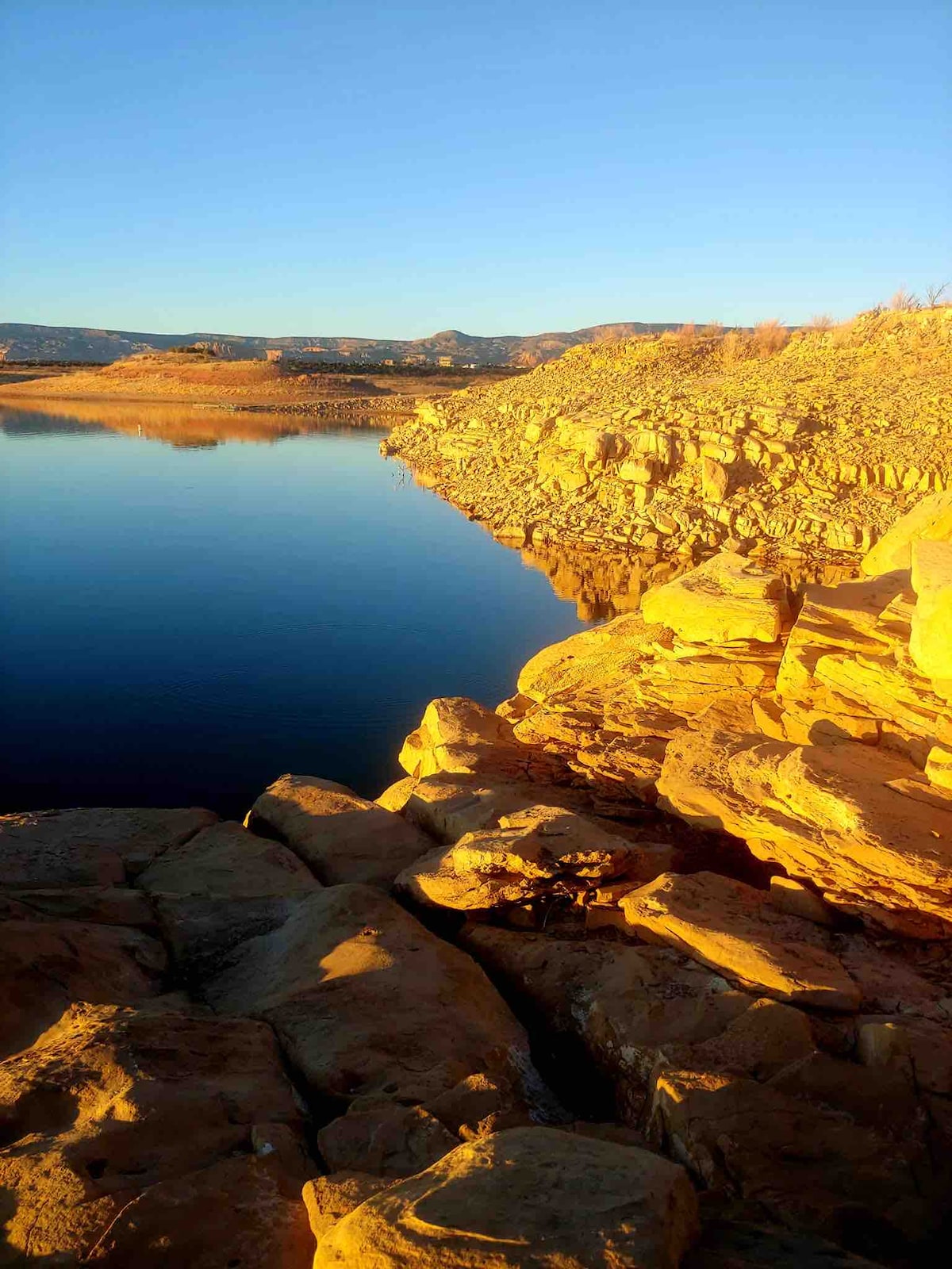 The shoreline of the Chama River is visible, featuring smooth rocks and warm, golden hues from the sunlight. Calm water reflects the surrounding landscape, which includes distant hills under a clear blue sky.