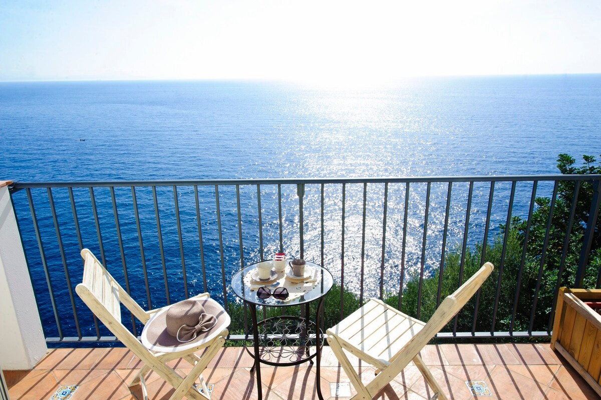 A private balcony is positioned over the shimmering sea, featuring two wooden chairs and a small round table. Sunlight creates a bright atmosphere, enhancing the tranquil view of the water extending to the horizon.