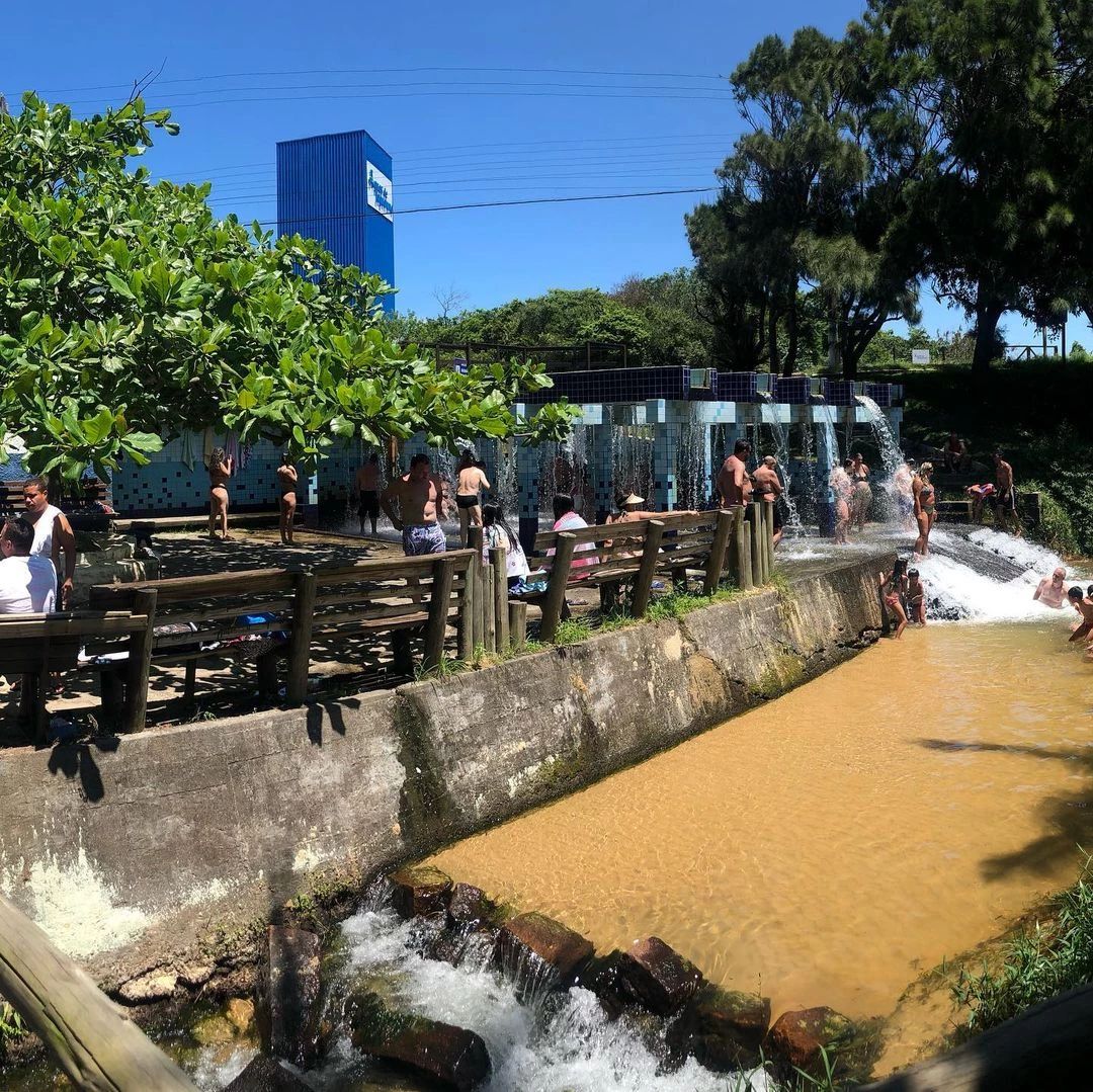 A vibrant outdoor area is surrounded by lush greenery and features several wooden picnic tables. A gentle water flow is visible beside a sandy path, leading to a playful water slide area where guests are enjoying leisure time.