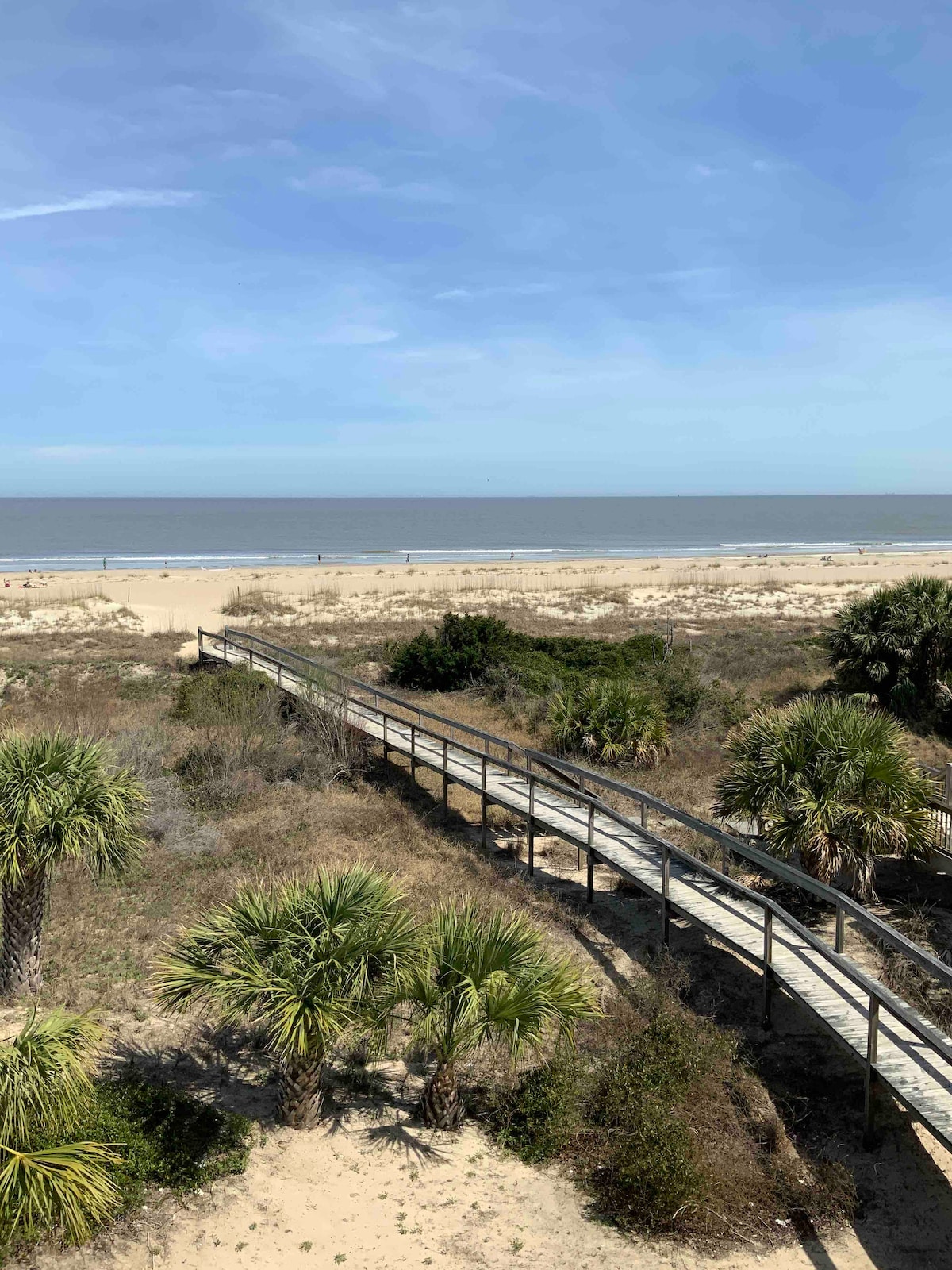 A sandy beach is visible, featuring a wooden walkway leading to the shoreline. Sparse vegetation, including palm trees, borders the path. The tranquil ocean stretches out in the distance under a clear blue sky, enhancing the natural coastal setting.