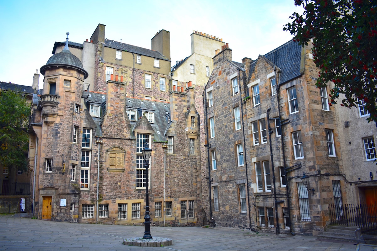 A historic building facade is displayed, featuring a mix of stone walls and architectural elements, with large windows and a tower. The courtyard includes a lamppost and is framed by surrounding structures, showcasing the character of the central Edinburgh area.