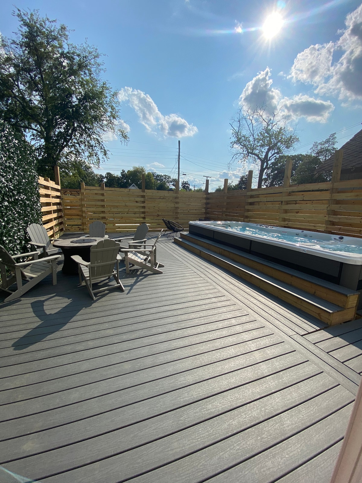 An outdoor space features a sleek hot tub situated beside a wooden deck. Surrounding the area are natural wood paneling and lush greenery, providing privacy. Comfortable seating is arranged around a table, and sunlight enhances the inviting atmosphere.