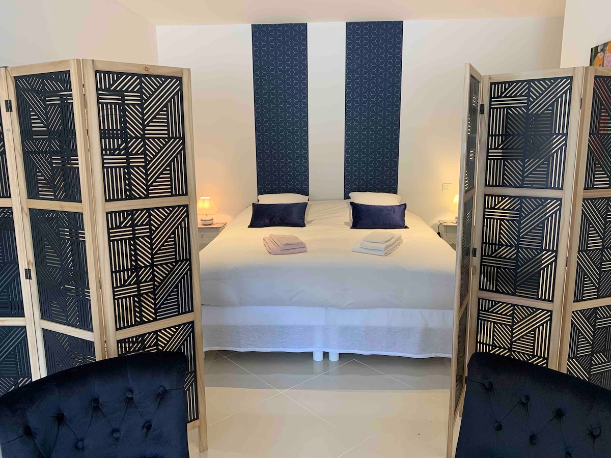 A spacious sleeping area is defined by wooden folding screens adorned with a geometric pattern. A large bed is presented with crisp white linens and two neatly arranged towels. Soft bedside lamps provide gentle illumination on either side.