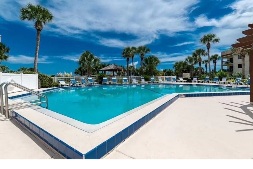 A spacious heated pool is surrounded by palm trees and lounge chairs, offering ample seating. A shaded pavilion adds to the inviting atmosphere, while clear blue skies and fluffy clouds create a serene backdrop for relaxation. The area features tiled edges and a clean, well-maintained appearance.
