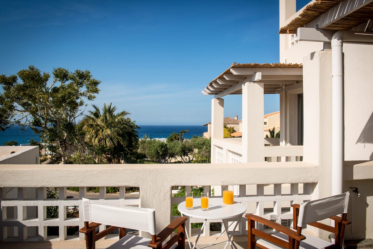 A serene balcony setting features a small round table with two orange drinks and two white chairs, overlooking lush greenery and the blue sea. Sunlight brightens the scene, highlighting the spacious outdoor area of the villa.