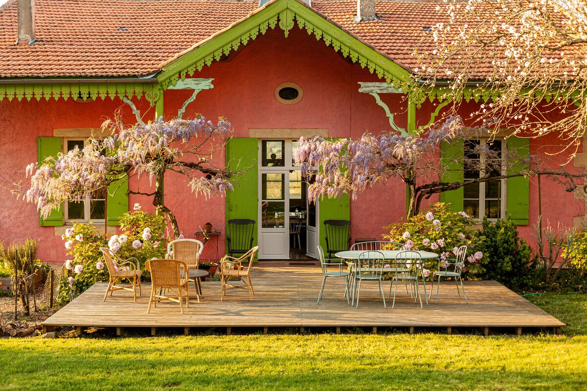 A vibrant house with pink walls is complemented by green shutters, creating a cheerful exterior. The wooden terrace features a variety of seating options surrounded by blooming flowers, with lush greenery visible. Two large trees provide shade, enhancing the outdoor space ideal for relaxation.